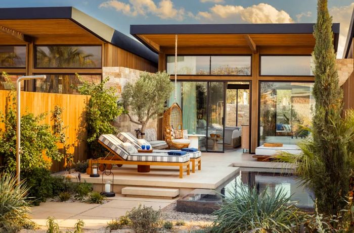 A patio adjoining a wooden bungalow featuring glass walls, encircled by Mediterranean flora and two striped lounge chairs.