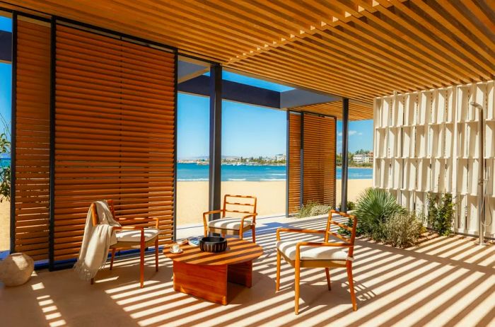 A terrace features three wooden chairs gathered around a table, complemented by a white architectural screen in the background and an outdoor shower, all playfully shaded by stripes of sunlight.