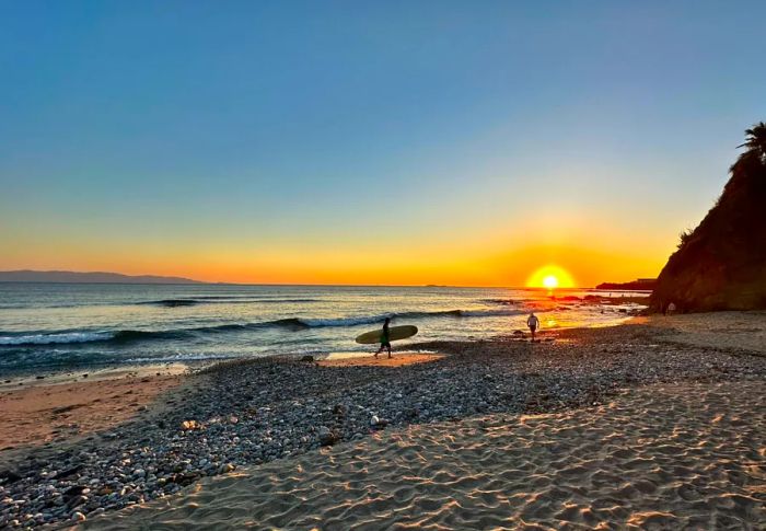 A person walking along the beach at sunset with a surfboard in hand