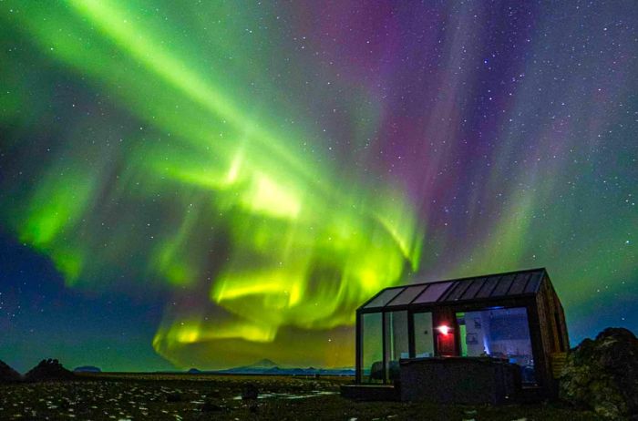 Bed inside a glass house in Iceland beneath vibrant green and purple northern lights.