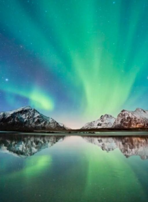 A sky mirrored in water, adorned with green ribbons of the aurora borealis, or northern lights, over Lofoten in Norway.