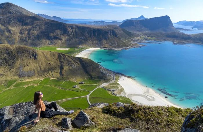 A woman perched on a high cliff overlooking Hauklandstranden beach in Lofoten, Norway