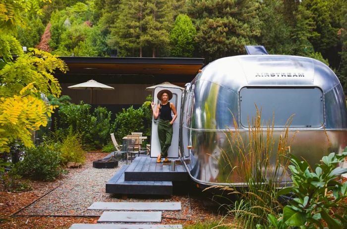 A person stands outside an Airstream, surrounded by trees in the background.