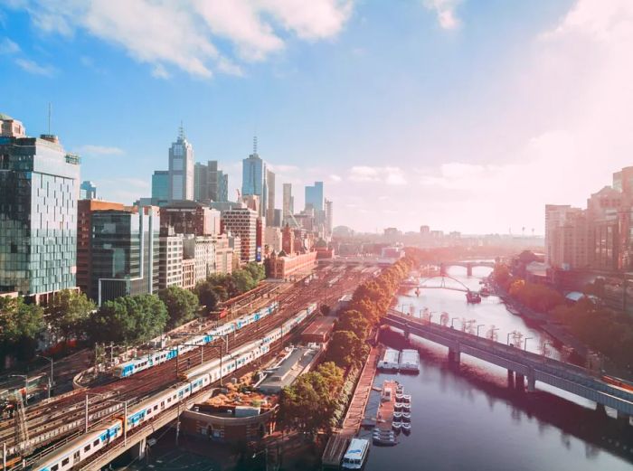 A panoramic view of Melbourne showcasing skyscrapers on the left, with trains and tracks alongside the waterfront on the right.