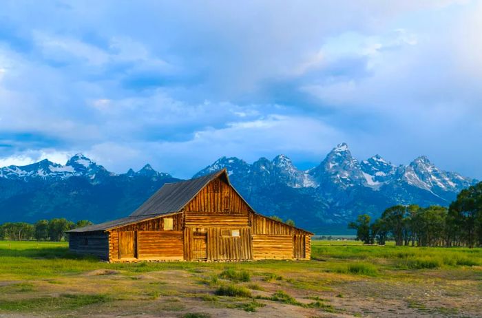 A log cabin is set against a grassy field with a stunning mountain range in the background near Jackson, Wyoming.