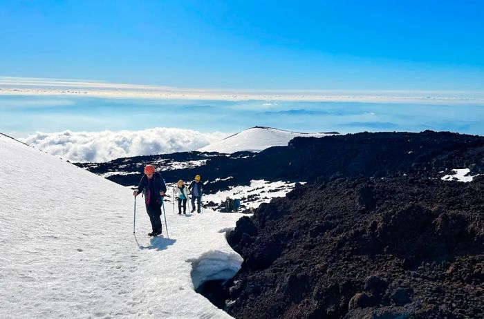 Hikers wielding ski poles trek through snow beside dark volcanic ash.