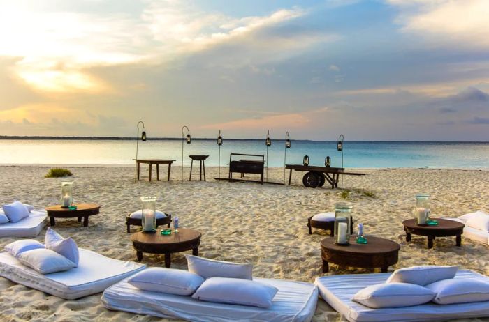 A cozy setup with cushions and low tables on the beach of Mnemba Island in Zanzibar, Tanzania, offers a picturesque view of a slightly cloudy sunset sky.