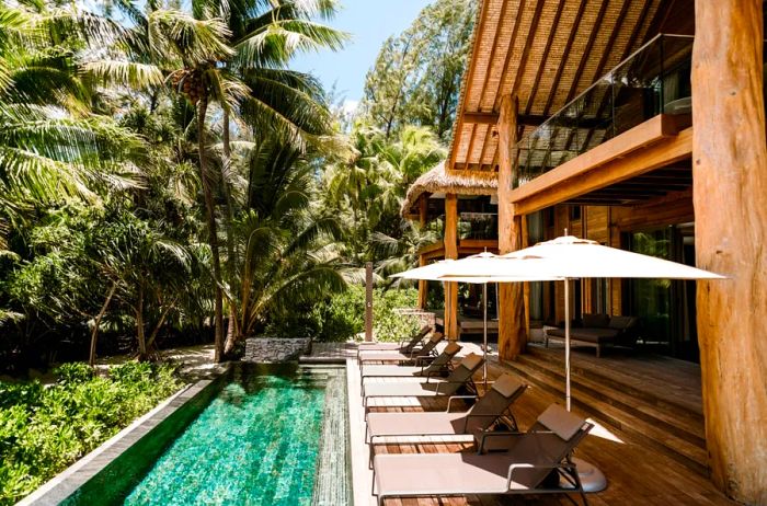 The exterior of a villa at The Brando features a pool, lounge chairs, white umbrellas, and swaying palm trees.