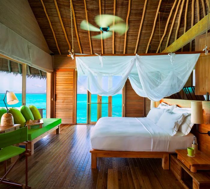 The interior of a Water Villa at Six Senses Laamu in the Maldives features a thatched roof, a bed with a mosquito net, and views of the turquoise waters that surround the villa.