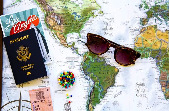 A pair of sunglasses, straight pins, a Los Angeles brochure, and a passport set against a world map.