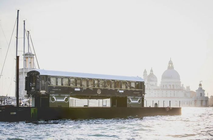 A train car is positioned on a barge with a cathedral in the background, set against the beautiful backdrop of Venice.