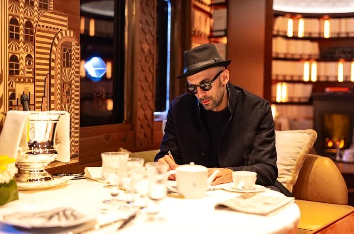 A man sporting dark sunglasses and a fedora is seen writing at a table adorned with drinking glasses and a ceramic teapot and cup, set against an intricately decorated wall.