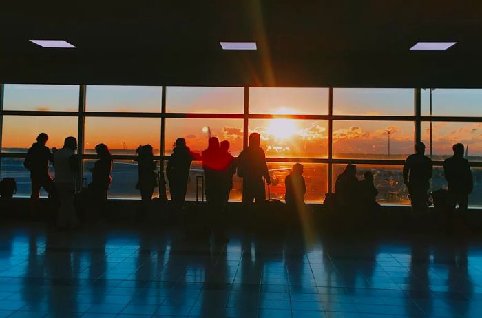 a group of travelers gazing out large airport windows at sunset