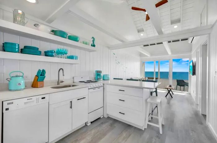 A bright white kitchen accented with turquoise details, including a teapot, dishware, and a coffee maker, all with a view of the ocean in the background.