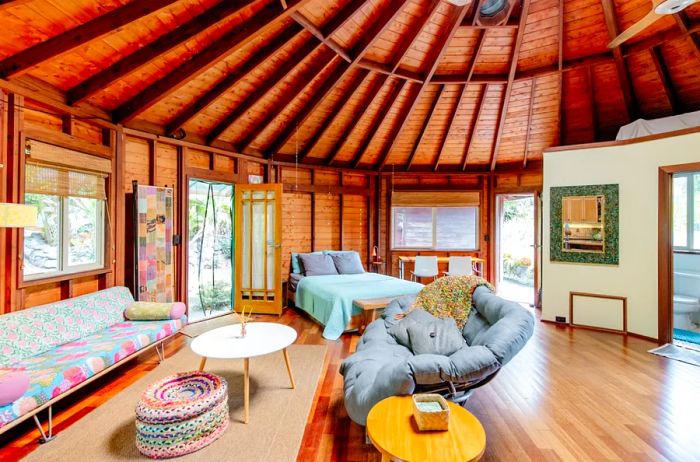 Interior of a unique 12-sided house featuring wooden ceilings, a patterned couch, a bed, and a cushioned chair.