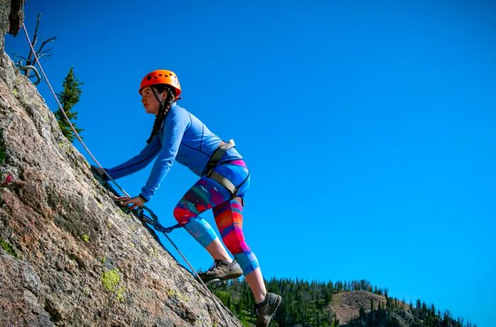 An individual dressed in vibrant climbing gear and a helmet scales a rock face at a steep angle.