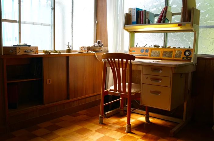 A quaint wooden desk and chair beside a cabinet and a bookshelf, situated in a corner room with windows on both sides