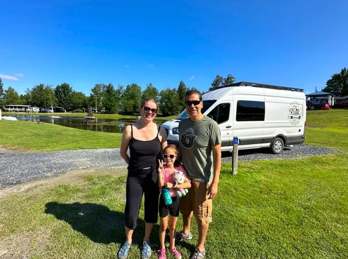 A woman, a child, and a man, all sporting sunglasses, pose in front of a white camper van in an outdoor setting.