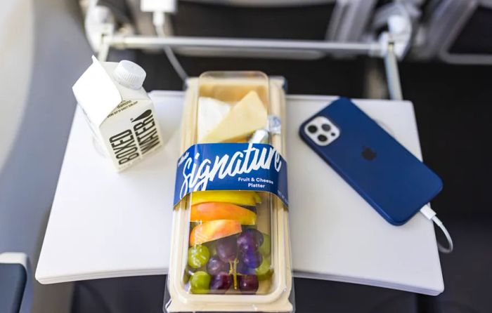 Alaska Airlines' Signature fruit and cheese platter elegantly arranged on an airplane tray alongside a small water bottle and a blue iPhone charging.