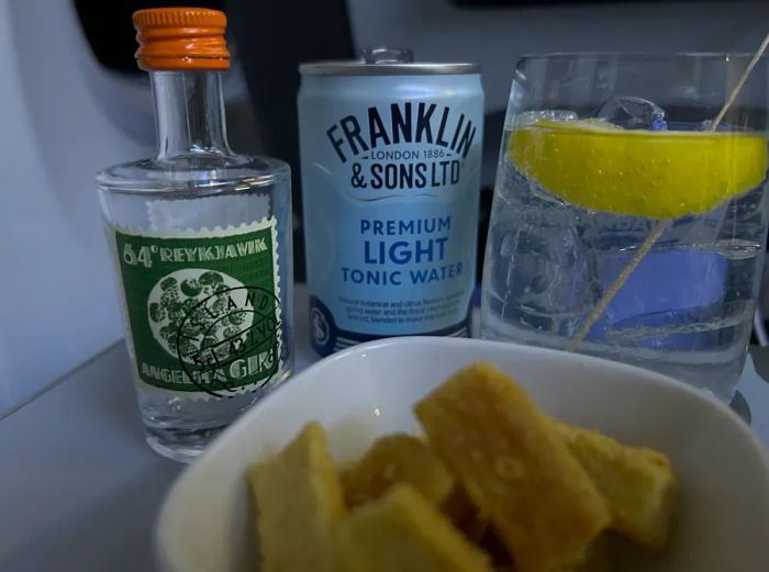 A refreshing Icelandic gin and tonic on Icelandair, showcasing a mini bottle of gin and a can of tonic alongside a glass garnished with a lemon slice and a small bowl of cheese crackers.