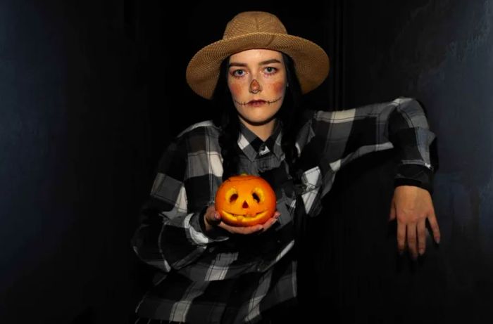 A girl dressed as a scarecrow holds a pumpkin in her hands on Halloween.