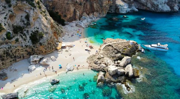 A picturesque white-sand beach alcove framed by towering rocks, boulders, and cliffs, with sunbathers lounging on the shore and a boat gliding across the turquoise waters of Sardinia, Italy