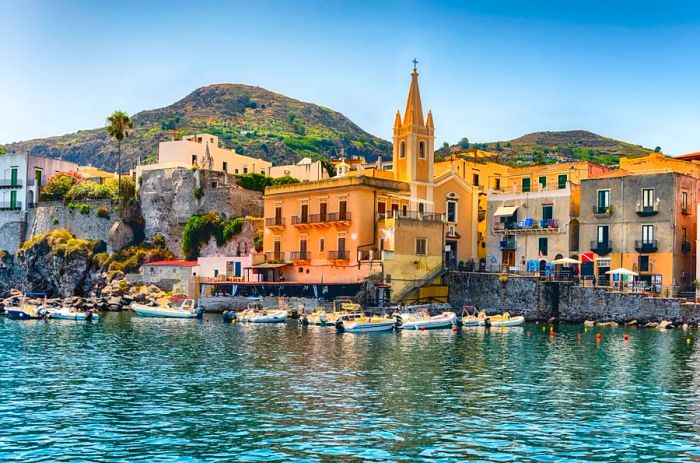 View of a vibrant coastal village in Lipari, the largest of the Aeolian Islands, featuring a pink and tan building/church with a steeple in the foreground and small boats moored nearby