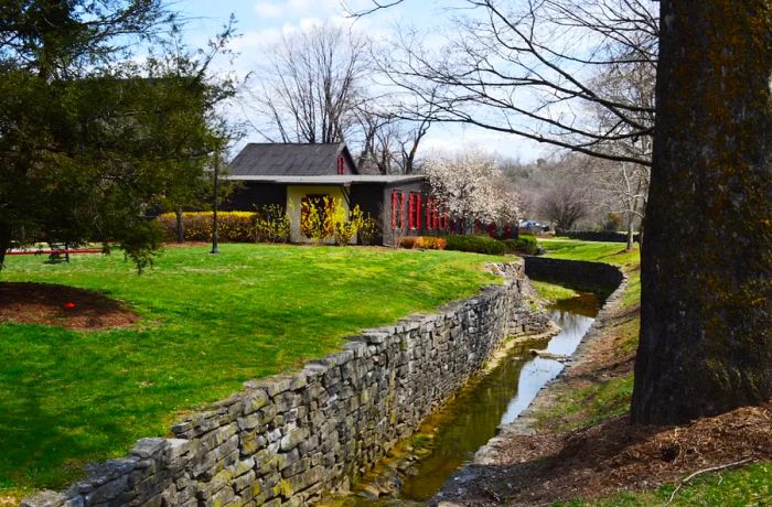 A gray house adorned with red window shutters stands beside a small stream bordered by stone walls.