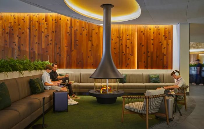 A man and two teenagers relax in an airport lounge, featuring banquette seating, wood paneling, and a central round firepit.