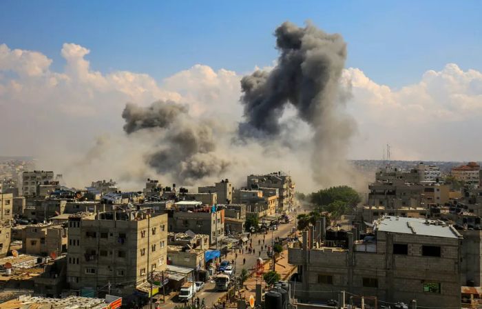 A distant view captures Israeli air strikes in Gaza, featuring bombed structures and dark gray plumes of smoke.