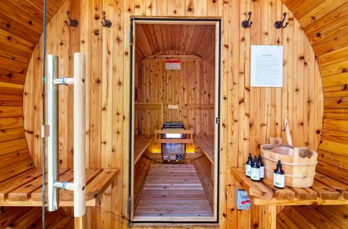 An open wooden barrel sauna, featuring a small table with a wooden bucket, brush, and four small bottles nearby