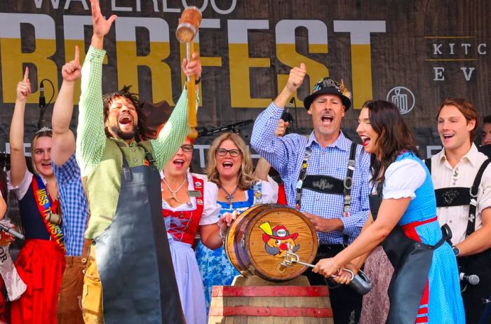 Celebrants in traditional German outfits tapping a keg featuring a whimsical cartoon logo.