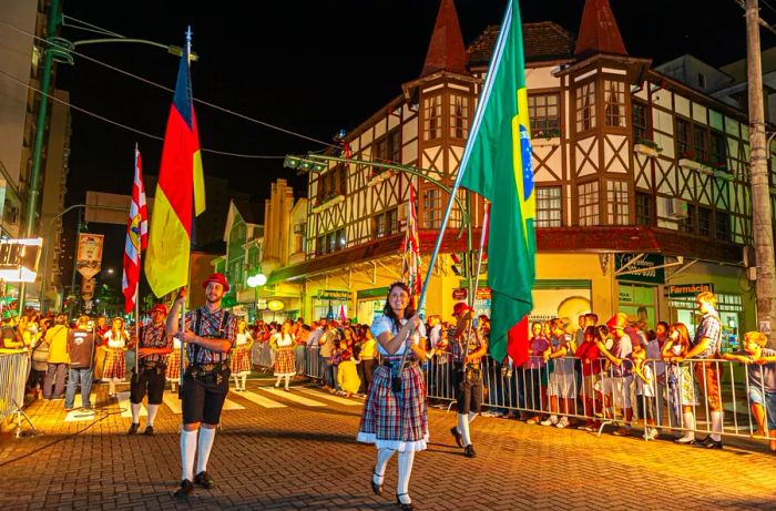 Festive participants in dirndls and lederhosen wave Brazilian and German flags during a nighttime parade.