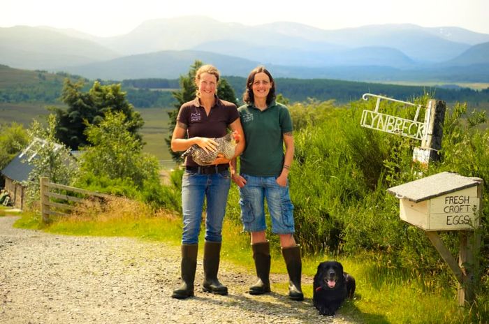 Lynn Cassells and Sandra Baer pose with a chicken and dog beside the Lynbreck Croft sign.