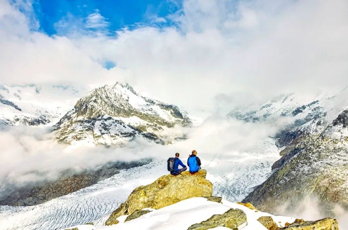 Two individuals seated on a rock, gazing out over snow-capped mountains and drifting low clouds.