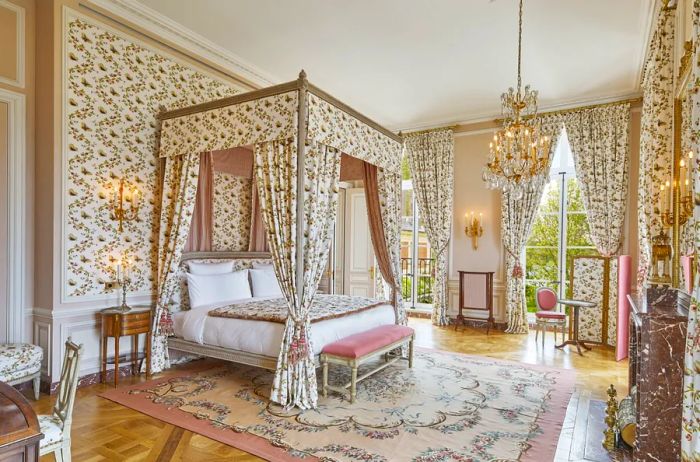 A lavish hotel room (predominantly white and gold) at the luxurious Palace of Versailles hotel features patterned wallpaper, curtained bedposts, and antique decor reminiscent of Marie Antoinette's style.