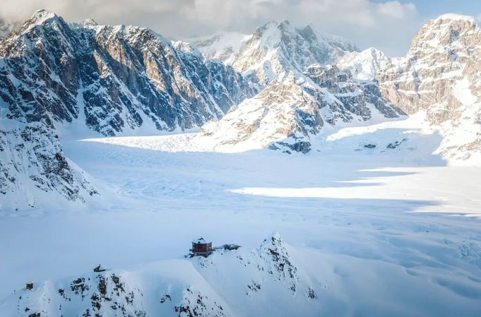 Aerial view of the cozy Sheldon Chalet perched on Ruth Glacier, enveloped by snow and towering mountains.