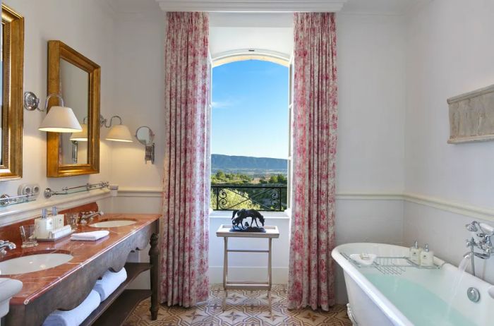 The spacious bathrooms at La Bastide de Gordes feature double sinks, a classic white clawfoot tub, and picturesque views of the Provencal countryside.