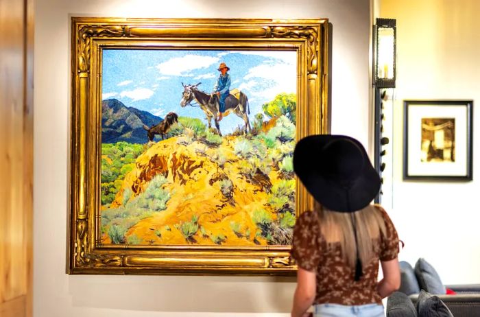 A woman wearing a hat admires a painting of a man on a donkey in the desert, part of The Blake Hotel's art collection.