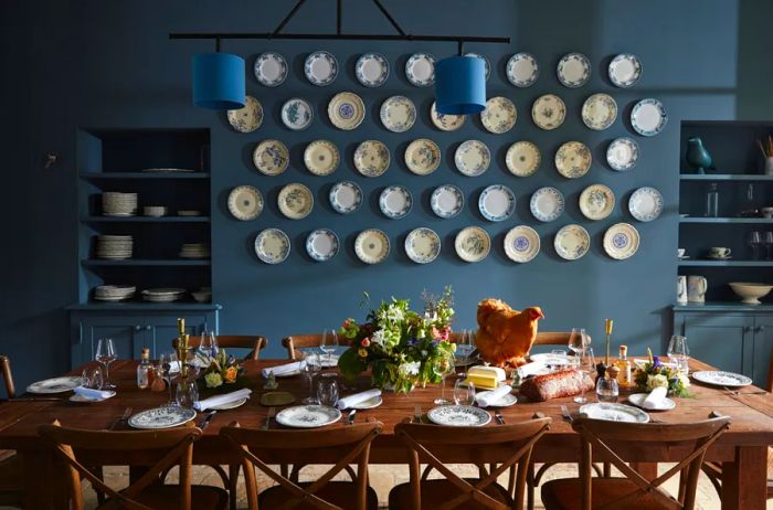 A large wooden dining table set for a meal, showcasing approximately 50 decorative plates displayed against a gray wall at Primard.