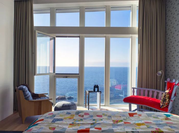 This guest room at Fogo Island Inn showcases a vibrant red cushioned chair and expansive floor-to-ceiling windows that offer stunning ocean views.