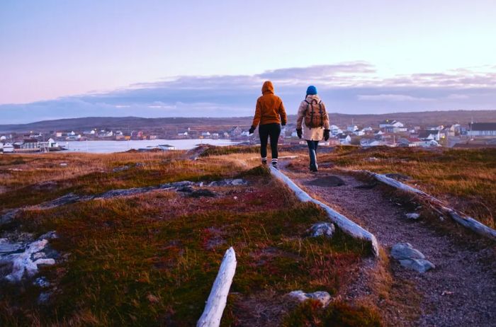 Two hikers traverse Fogo Island, with a cluster of homes visible in the background.