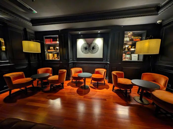 A stylish bar featuring orange velvet chairs, small black tables, wood flooring, and dark walls adorned with art.