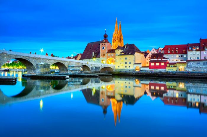 Germany, Bavaria, Upper Palatinate, Regensburg - The Stone Bridge, St. Peter's Church, and the Old Town of Regensburg reflecting on the Danube River at night.