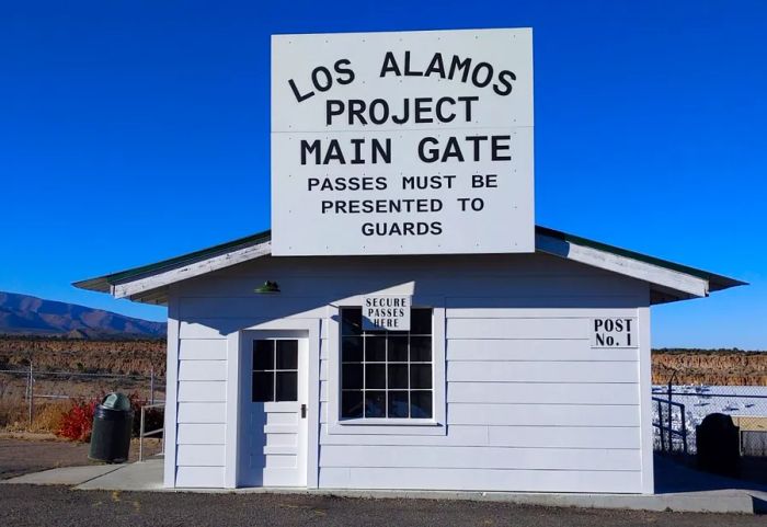 This is a replica of the security gate from the Manhattan Project, a quaint white wooden structure topped with a prominent sign reading 'Los Alamos Project Main Gate.'