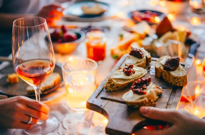 A hand holding a wine glass of rosé alongside another hand with a small wooden cutting board featuring appetizers.