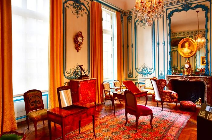 An elegant recreation of an 18th-century salon at the Musée Carnavalet, featuring gold drapes along towering windows and a grand mirror.