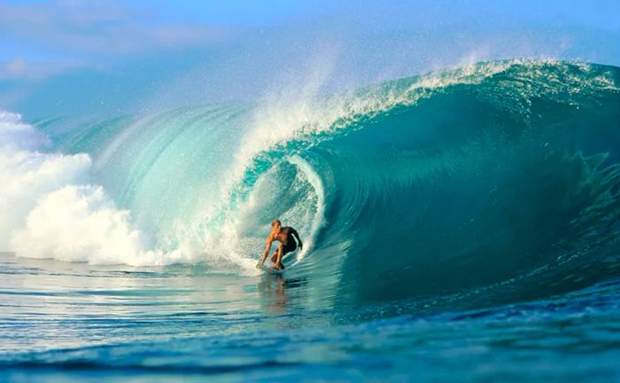 A surfer rides a vibrant blue wave under sunny skies.