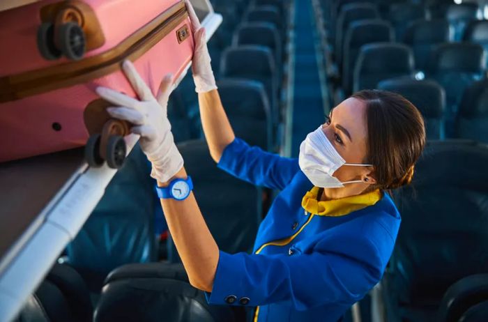 A masked and gloved flight attendant placing a pink carry-on roller bag into the overhead compartment