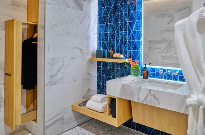 A Delta One lounge shower room adorned with a sink, blue tiles, and white-and-gray marble, complete with a robe hanging in the foreground and a pocket door closet for ironing services.